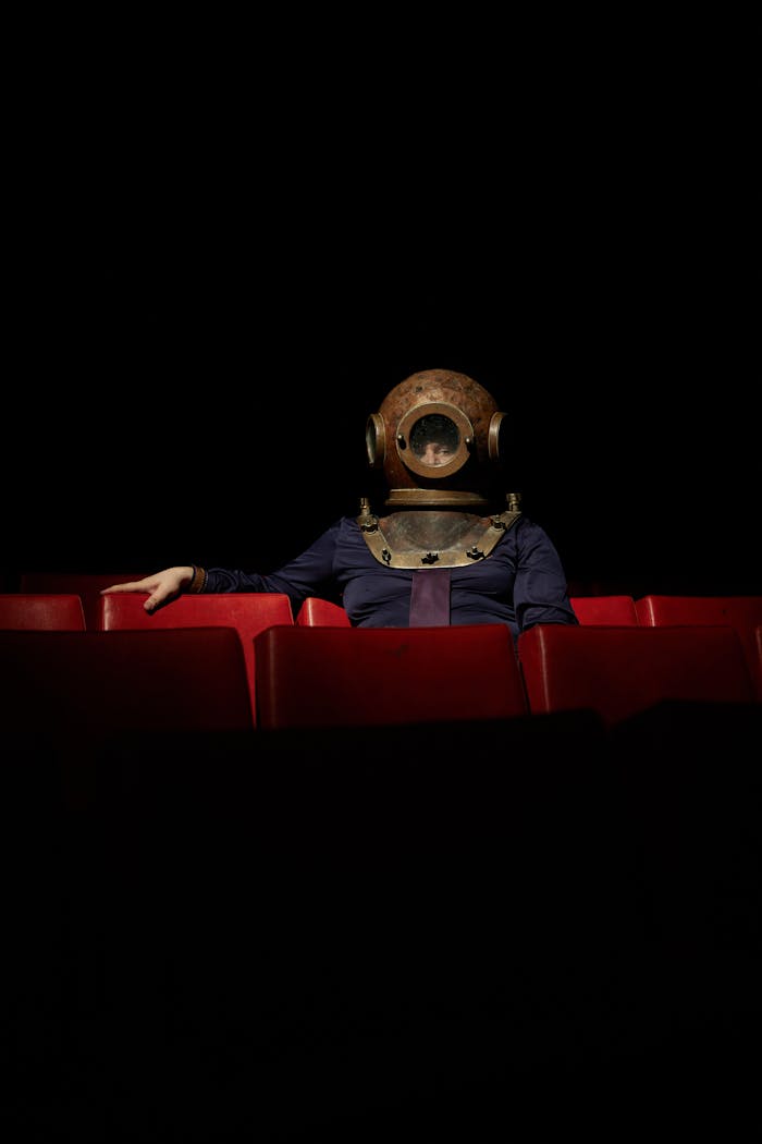 Person wearing vintage dive helmet in dark theater with red seats.